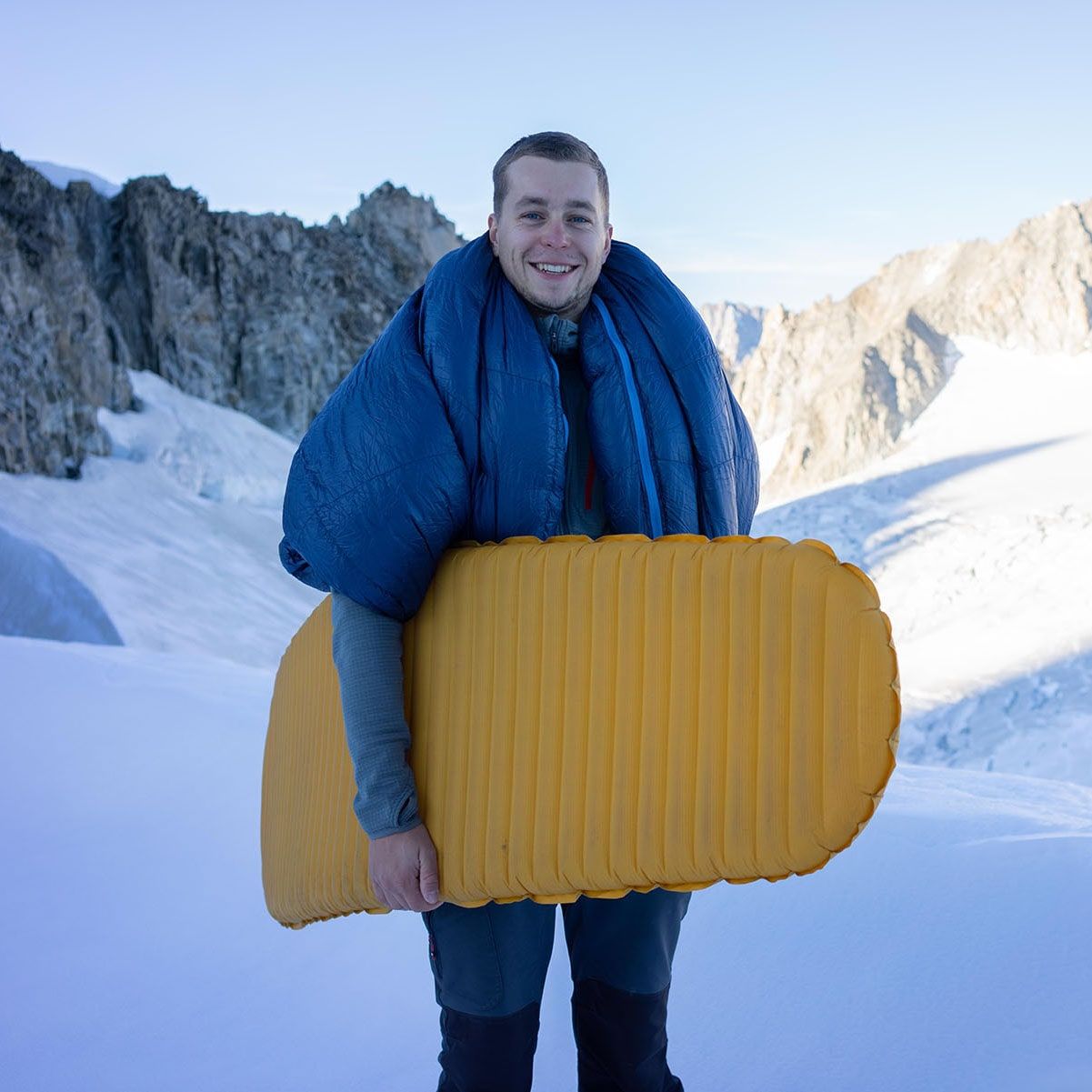 Osoba držící nafukovací žlutou karimatku a kolem krku přehozený ultralehký péřový spacák