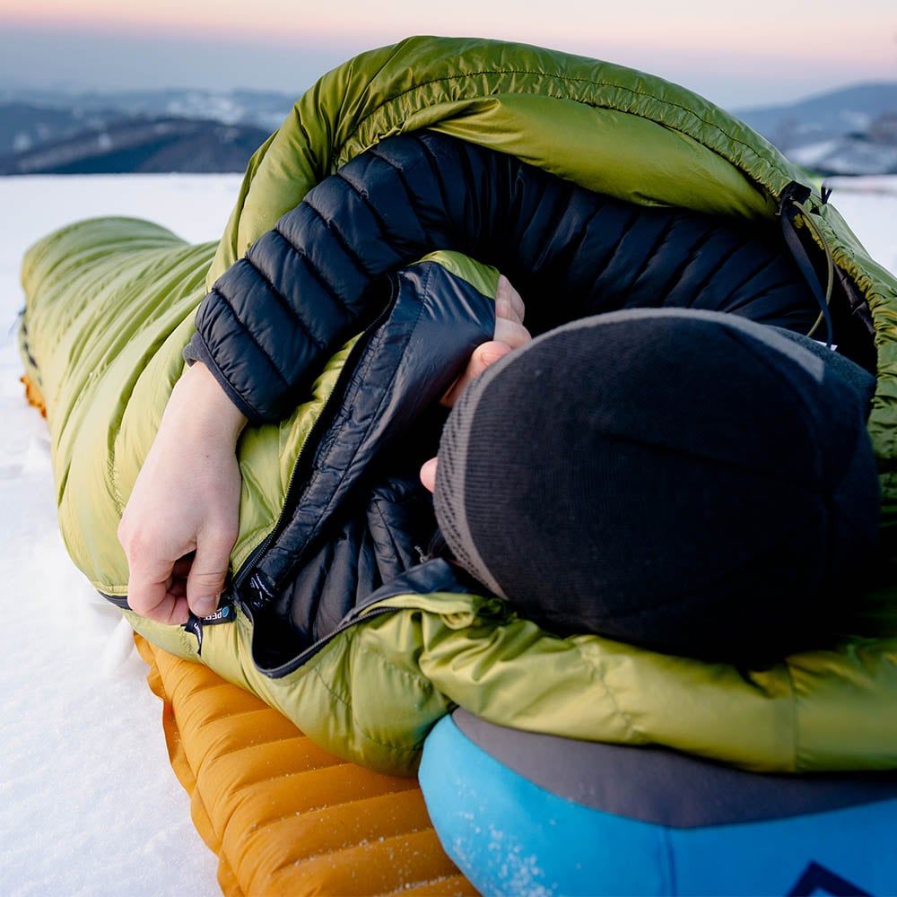 Osoba ležící ve spacáku Cumulus Lite Line 400 v zelené barvě při kempování na sněhu na nafukovací karimatce a polštářku.
